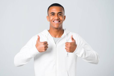 Genç adam koyu tenli bir başparmak yukarıya hareket veren ve çünkü gülümseyen beyaz kazak ile gri arka plan üzerinde başarı elde etti