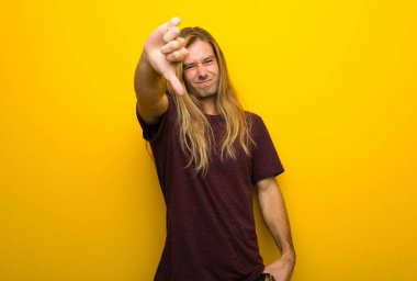 Başparmak aşağı negatif ifade işaretiyle gösterilen sarı duvarın üzerinden uzun saçlı sarışın adam