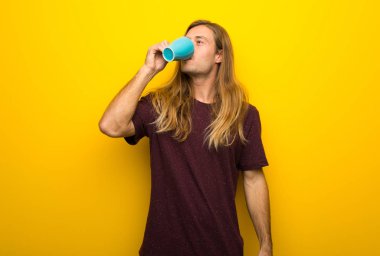 Bir fincan sıcak kahve tutan sarı duvarın üzerinden uzun saçlı sarışın adam