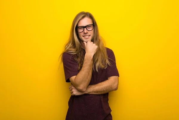 Blond man with long hair over yellow wall with glasses and smiling