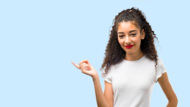 Young girl with curly hair pointing finger to the side and presenting a product while smiling in a confident pose on blue background