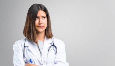 Doctor woman with stethoscope having doubts and with confuse face expression while bites lip. Questioning an idea on grey background