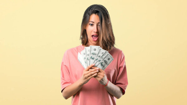 Surprised Young girl with pink shirt taking a lot of money on yellow background
