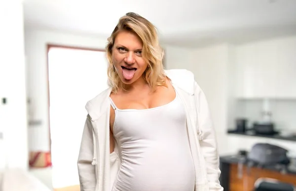 怀孕的金发碧眼的女人与白色运动衫显示舌头在镜头有有趣的外观在她的房子 — 图库照片