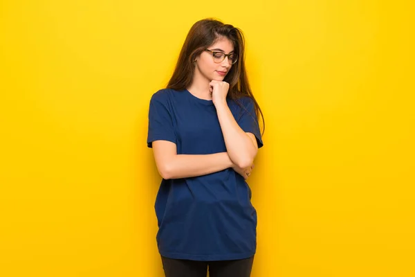 Young woman with glasses over yellow wall looking down with the hand on the chin