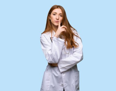 Kızıl saçlı doktor kadın parmağını ağzına izole mavi zemin üzerine koyarak sessizlik jest belirtisi gösteren