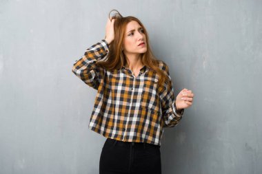 Young redhead girl over grunge wall having doubts while scratching head