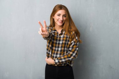 Genç Kızıl saçlı kız gülümseyerek ve zafer işaret gösteren grunge duvarın üzerinden