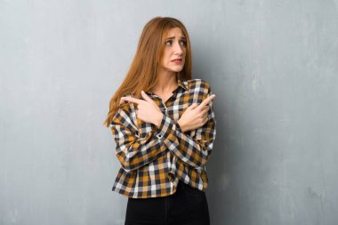 Young redhead girl over grunge wall pointing to the laterals having doubts