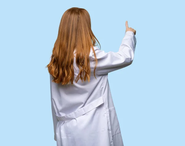 Руда Жінка Лікар Вказує Назад Індексним Пальцем Ізольованому Синьому Фоні — стокове фото