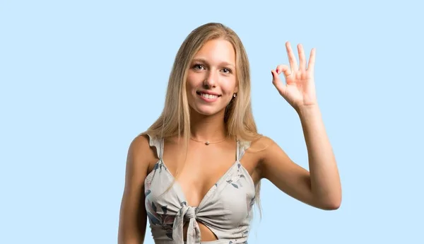年轻的金发碧眼的女人用手指显示一个确定的标志 在蓝色背景下面对幸福和满足 — 图库照片