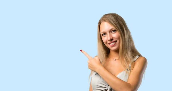 年轻的金发碧眼的女人用手指指着身边展示一个产品或一个想法 同时期待着在蓝色背景上微笑 — 图库照片