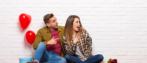 Couple in valentine day at indoors pointing finger to the side with a surprised face
