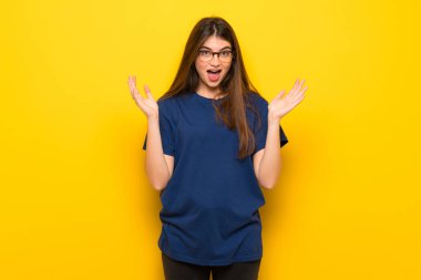 Sürpriz ve şok yüz ifadesi ile sarı duvarın üzerinden gözlüklü genç kadın