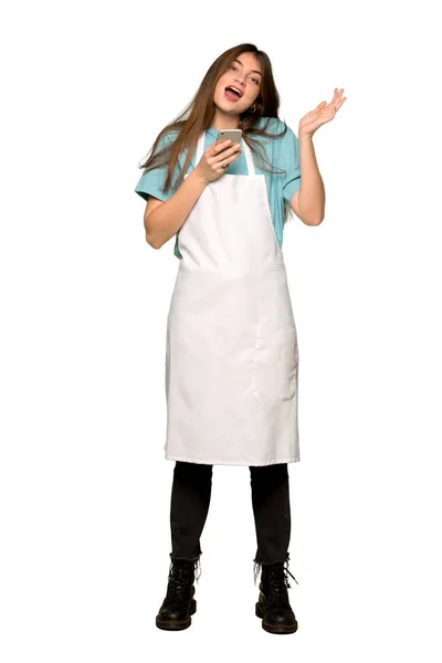 Full-length shot of Girl with apron surprised while sending a message with the mobile on isolated white background