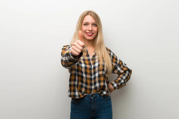 Blonde young girl over white wall giving a thumbs up gesture because something good has happened