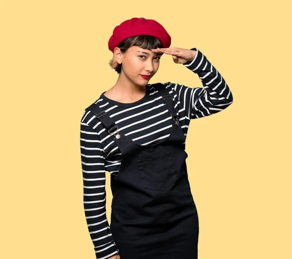 Young woman with beret looking far away with hand to look something over yellow background