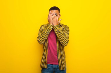 Gözleri eller tarafından kapsayan ve parmaklarının arasından seyir sarı zemin üzerine genç afro Amerikalı adam