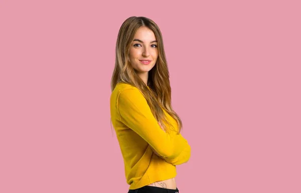 年轻的女孩与黄色毛衣保持手臂交叉在侧向位置 而微笑在孤立的粉红色背景 — 图库照片