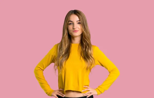 年轻的女孩与黄色毛衣摆姿势与手臂在臀部和笑看在前面在孤立的粉红色的背景 — 图库照片