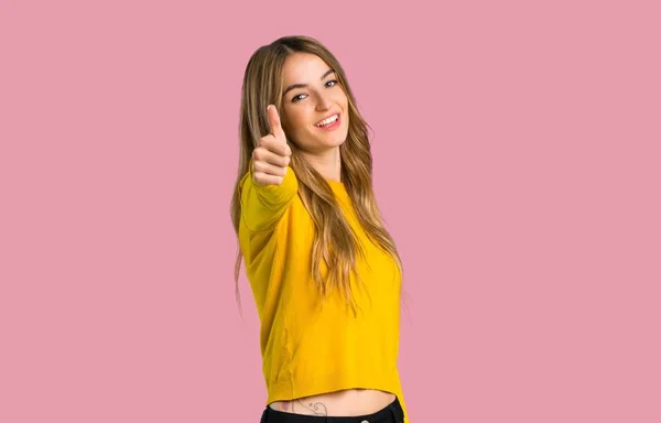 年轻的女孩与黄色毛衣竖起大拇指的姿态 因为一些好的事情发生在孤立的粉红色背景 — 图库照片