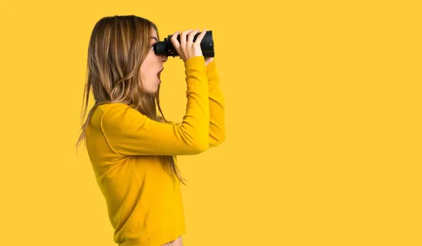 黄色いセーターと分離された黄色の背景に双眼鏡を使って遠くに見て若い女の子 — ストック写真