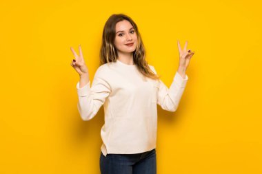 Genç kız gülümseyerek ve iki elini zafer işaretiyle gösterilen sarı duvarın üzerinden