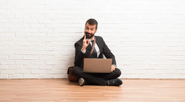 Businessman with his laptop sitting on the floor showing and lifting a finger in sign of the best