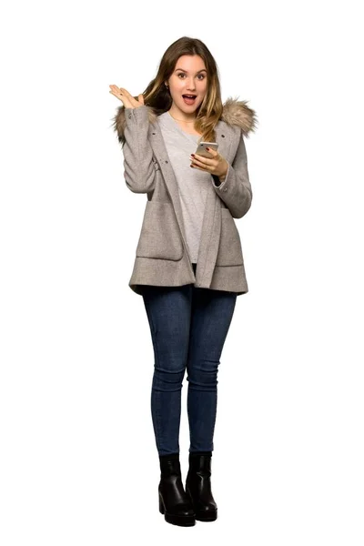 分離の白い背景の上 携帯電話でメッセージを送信中に驚きのコートで 代の女の子のフルレングス ショット — ストック写真