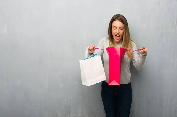 Young woman on textured wall surprised while holding a lot of shopping bags