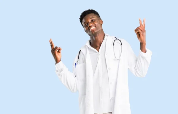 笑顔と分離の青い背景に陽気な顔と両方の手で勝利のサインを示すアフリカ系アメリカ人の医者 — ストック写真
