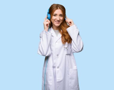 Redhead doctor woman listening to music with headphones on isolated blue background