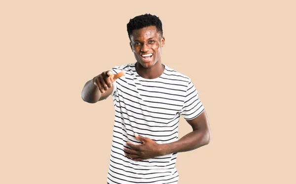 Dark skinned man with striped shirt pointing with finger at someone and laughing a lot on isolated ocher background