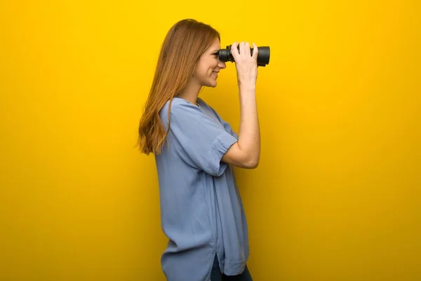 黄色の壁の背景との距離で双眼鏡で見て若い赤毛の女の子 — ストック写真