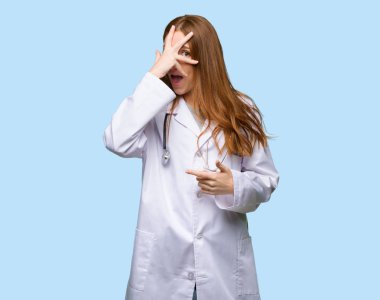 Kızıl saçlı Doktor kadınla sürpriz ve izole mavi zemin üzerine şok yüz ifadesi