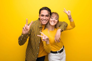 Canlı sarı arka plan üzerinde genç çift gülümseyerek ve her iki eliyle zafer işareti gösteren