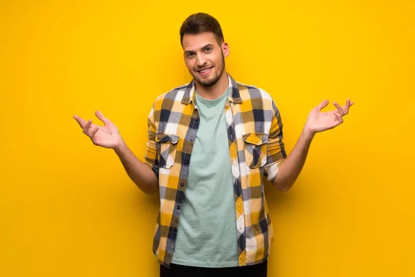 Handsome man over yellow wall having doubts while raising hands