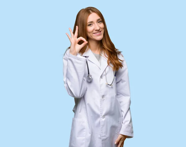红发医生妇女显示一个确定的标志与手指在孤立的蓝色背景 — 图库照片