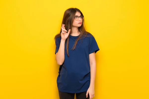 Young woman with glasses over yellow wall with fingers crossing and wishing the best