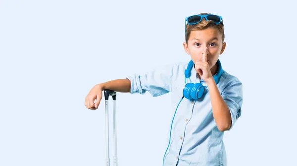 带着太阳镜和耳机的孩子带着他的行李箱旅行 在蓝色背景上露出闭上嘴和沉默的手势 — 图库照片