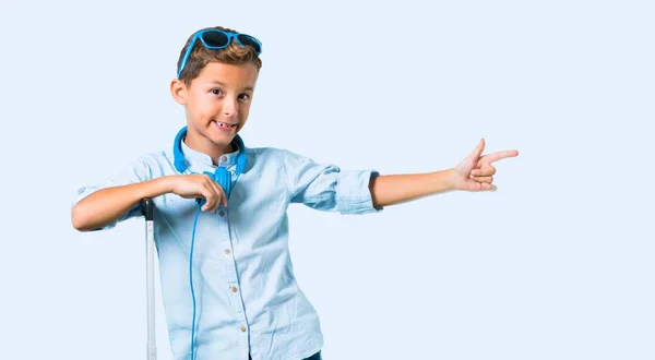 带着太阳镜和耳机旅行的孩子 他的手提箱用手指指着一边 并在蓝色背景上展示一个产品 — 图库照片