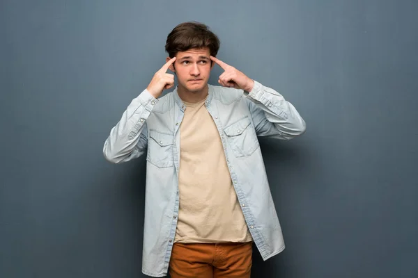 Teenager man with jean jacket over grey wall having doubts and thinking