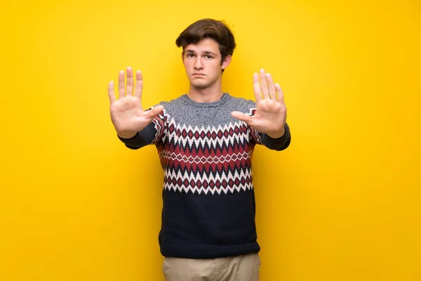 Teenager man over yellow wall making stop gesture and disappointed