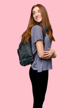 Kızıl saçlı öğrenci kadın izole pembe arka plan bir gülümseme ile omuz üzerinden arıyor