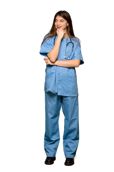 分離の白い背景の上のあごに手で側にいる若い看護婦さんのフルレングスのショット — ストック写真