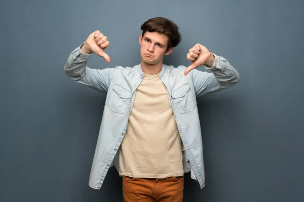 Teenager man with jean jacket over grey wall showing thumb down