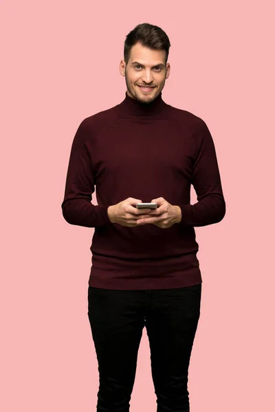 ピンクの背景の上 携帯電話でメッセージを送信するタートルネックのセーターを持つ男 — ストック写真