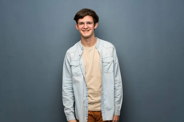 Teenager man with jean jacket over grey wall with glasses and happy