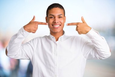 Beyaz gömlek odaklanmamış açık arka plan ağız ve yüz parmak ile işaret ederken mutlu ve keyifli bir ifade ile gülümseyen genç Afro-Amerikan adamla