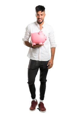 Beyaz gömlekli Arap bir gencin izole edilmiş beyaz arka planda büyük bir kumbara tutarken çekilmiş uzun bir resmi.